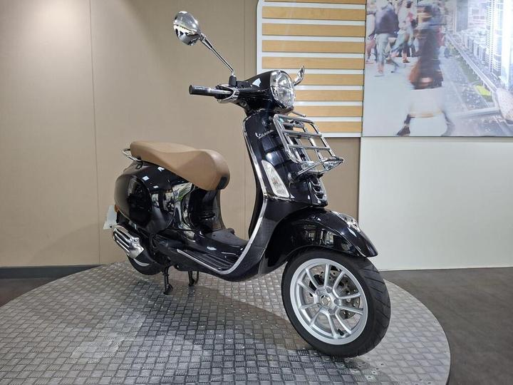 A black scooter with a brown seat is showcased on a circular display platform against a neutral background, featuring a modern urban image.