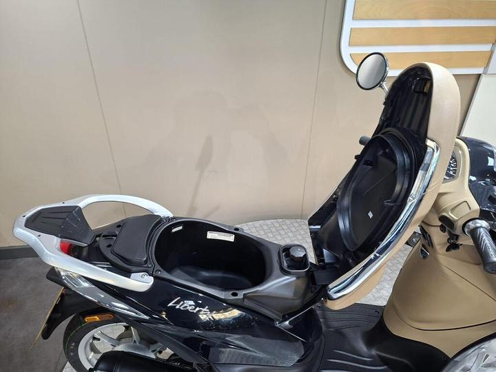 A close-up view of a black scooter's seat open, revealing a storage compartment underneath, with a seat design in cream and chrome accents. The background shows a simple, neutral wall.