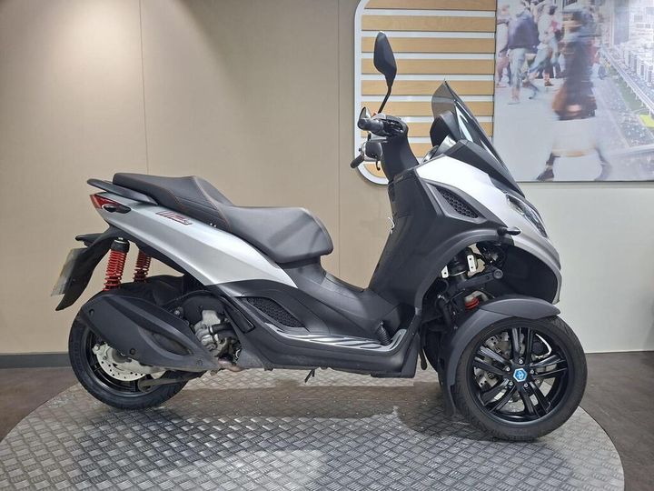 Piaggio MP3 300 HPE Sport (2022) A three-wheeled scooter in silver and black, featuring a prominent windshield, sleek design, and a red suspension system, displayed on a circular platform against a neutral wall background.