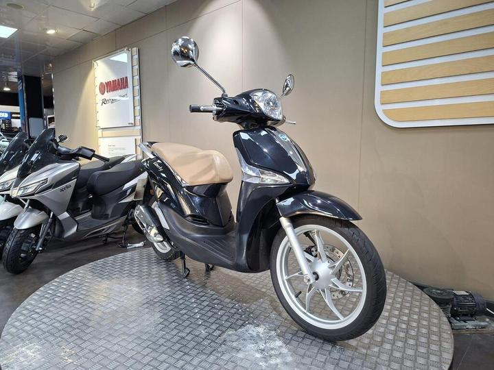 A black scooter with a light brown seat is prominently displayed on a circular metal platform in a showroom, surrounded by other motorcycles, including a silver Aprilia model. Behind the scooter, a Yamaha sign is visible on the wall.