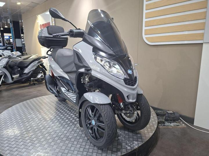 A gray three-wheeled scooter displayed on a circular platform in a showroom, featuring a windshield, storage compartment, and sporty design, with additional scooters visible in the background.