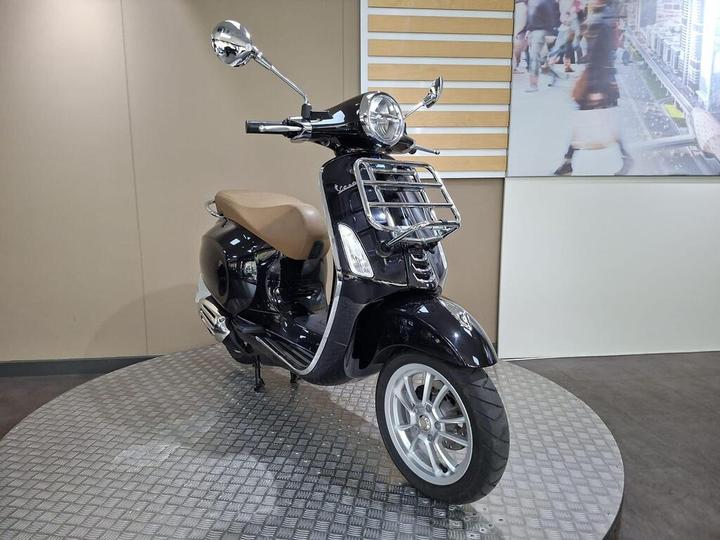 A sleek black scooter with a tan seat and chrome accents, positioned on a circular platform, set against a neutral background.