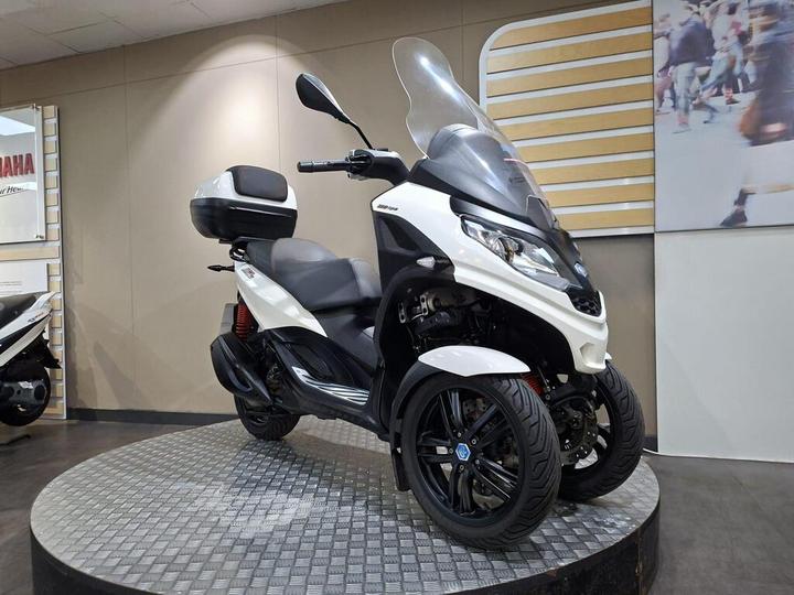 Three-wheeled scooter displayed on a circular platform in a showroom, featuring a sleek white and black design, a windshield, and a storage compartment on the back.