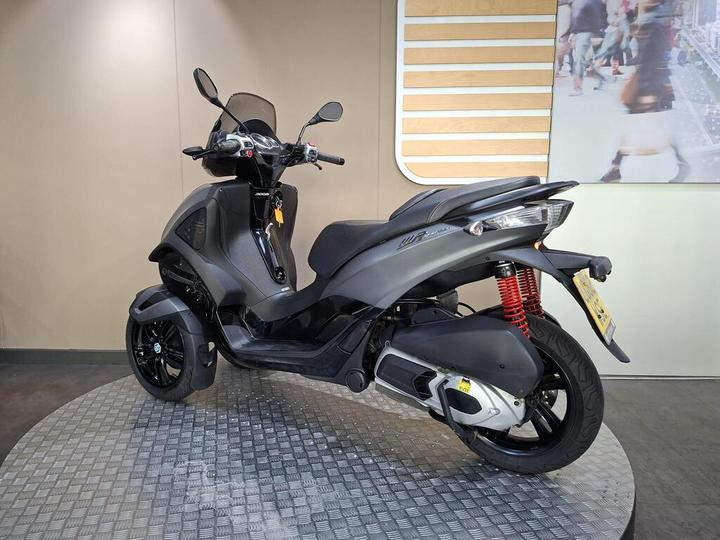 A black three-wheeled scooter displayed on a textured circular platform with a neutral background and a partially visible urban scene in the backdrop.