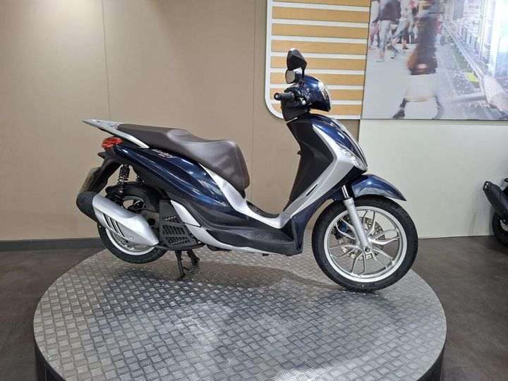 Piaggio Medley 125 2019 A blue and silver scooter parked on a circular platform, with a neutral-colored background and a blurred image of people walking in the distance.