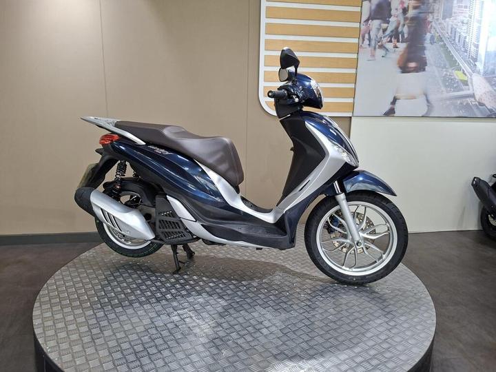 Piaggio Medley 125 2019 A blue and silver scooter parked on a circular platform, with a neutral-colored background and a blurred image of people walking in the distance.