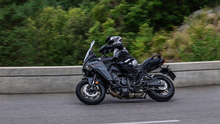 A rider in black and white motorcycle gear expertly maneuvers a sleek, black motorcycle along a curved mountain road, with greenery in the background.