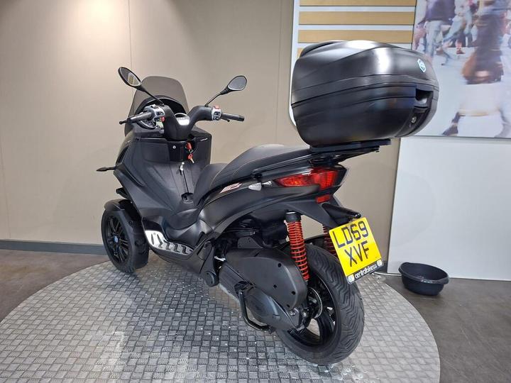 A black three-wheeled scooter with a rear storage box parked on a circular metallic platform, set against a neutral wall backdrop.