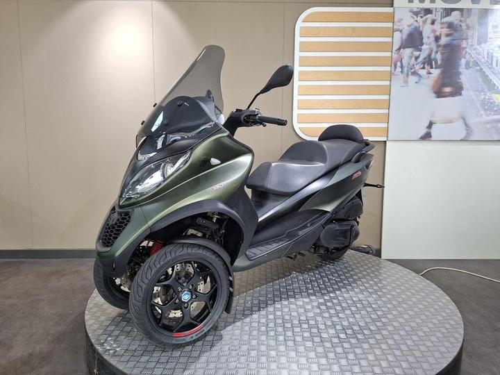 A green three-wheeled scooter displayed on a circular platform, featuring a front windshield, black seat, and alloy wheels, with a modern background showing a cityscape.