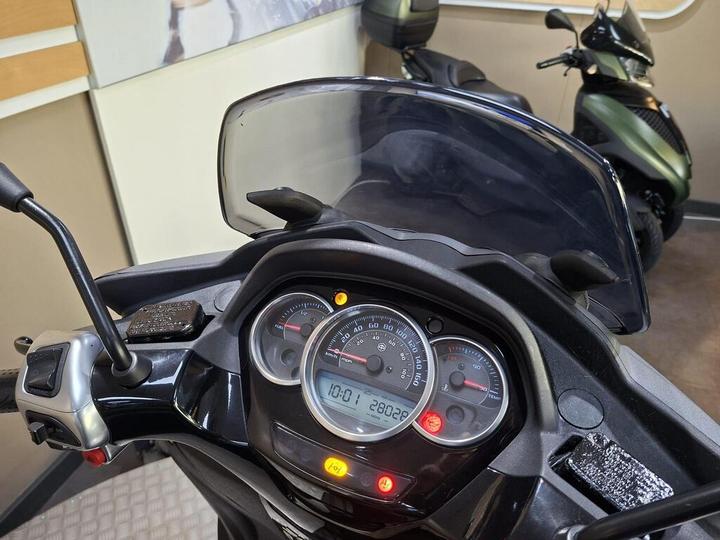 Close-up view of a scooter's dashboard displaying a speedometer and odometer, with various warning lights illuminated. In the background, two scooters are partially visible, along with a muted interior setting.