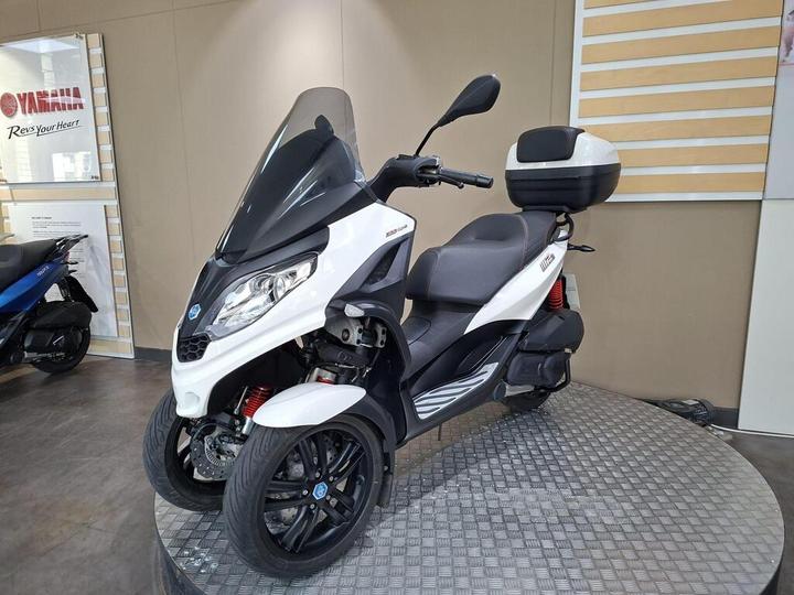 A three-wheeled scooter with a sleek white and black design, featuring a windshield and a rear storage box, displayed on a circular metallic platform. The background includes a Yamaha sign and informational displays.