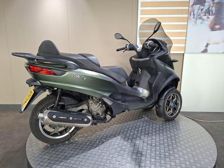 A green three-wheeled scooter displayed on a circular platform with a textured surface, featuring a black seat and a windscreen, against a plain wall background.