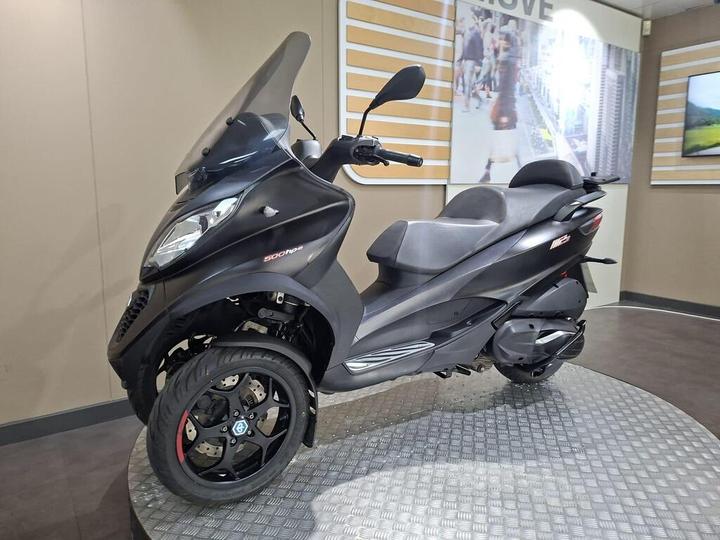 A black three-wheeled scooter displayed on a round metallic platform, with a transparent windscreen and a sleek design, set against a neutral background with subtle geometric patterns.