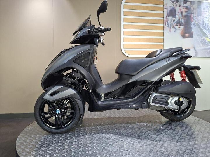 A sleek black scooter displayed on a circular platform, showcasing its modern design and features, with a backdrop of a minimalist exhibition space.