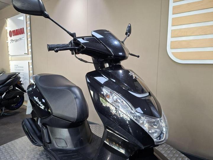 A black scooter is displayed in a showroom, featuring a sleek design with a clear windscreen, front lights, and a comfortable seat. The background includes a Yamaha logo and a glimpse of another scooter.