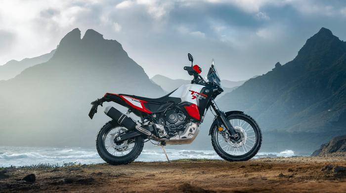 A motorcycle is parked on a rocky coastline with mountains in the background, under a partly cloudy sky.