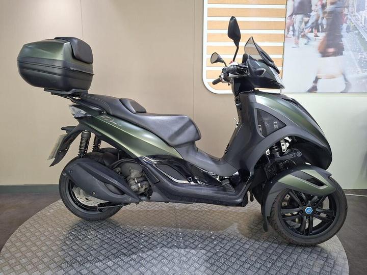 Piaggio MP3 300 Yourban LT 2018 A sleek black and green scooter displayed on a patterned surface, featuring a top case for storage, showcasing its modern design and dual front wheels for stability.