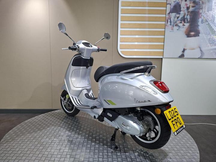 A silver Vespa scooter is displayed on a textured circular platform in an indoor showroom, with a modern backdrop featuring people and city imagery. The scooter is viewed from a three-quarter angle, highlighting its design and features.