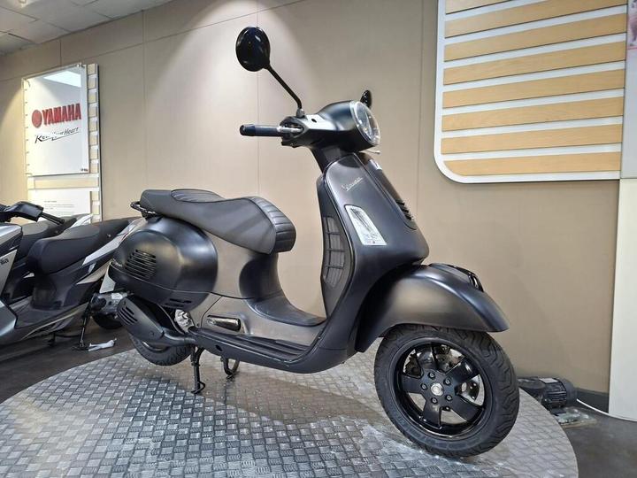 A sleek black scooter displayed on a textured floor, with modern design features and a prominent front headlight. The background shows a dealership setting with Yamaha branding.