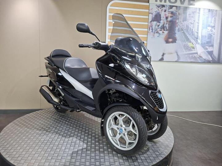 A black three-wheeled scooter displayed on a circular platform against a neutral backdrop, featuring a protective windshield and a sleek design.
