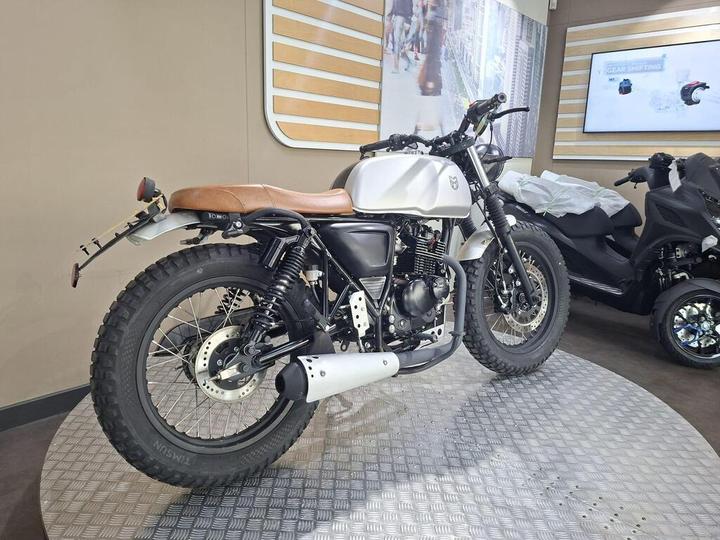 A custom motorcycle featuring a matte white fuel tank, brown leather seat, and rugged tires, displayed on a circular platform with a sleek background of wooden panels and digital screens.