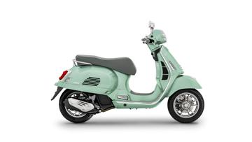 A light green Vespa scooter viewed from the side, featuring a gray seat, chrome accents, and a single headlight.