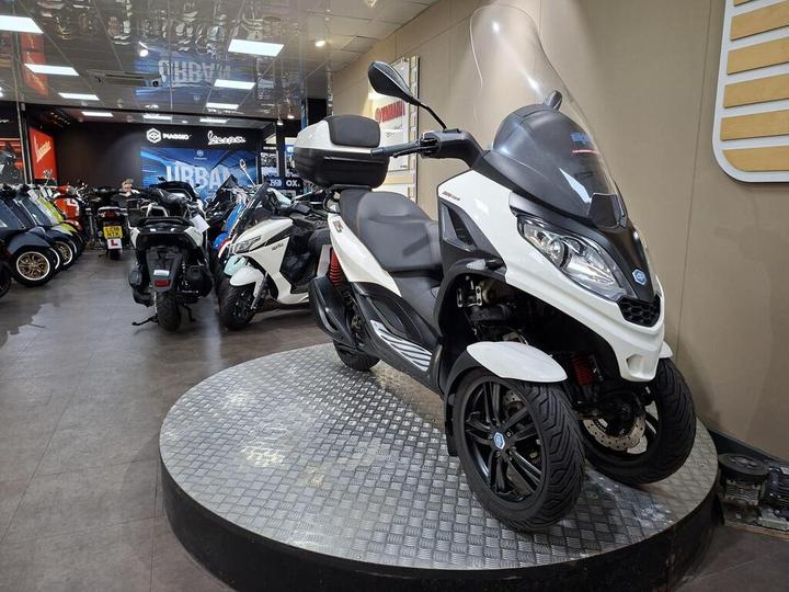 A white and black three-wheeled scooter displayed on a circular platform in a motorcycle showroom, with various other scooters and motorcycles in the background.