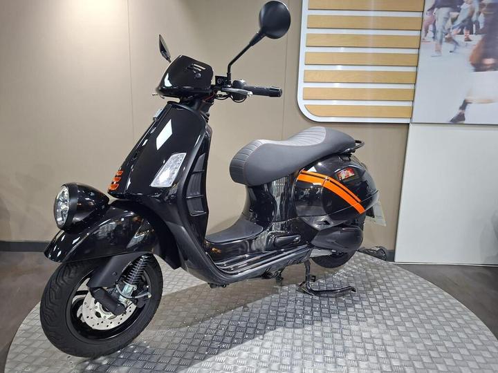 A sleek black scooter with orange accents is displayed on a checkered platform against a neutral background. The scooter features a modern design with a rounded headlight, a comfortable gray seat, and a visible rear wheel.