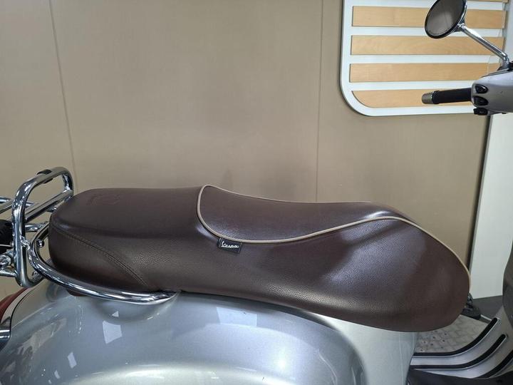 A close-up view of a brown motorcycle seat with a white piping detail, attached to a silver scooter, set against a neutral wall with wooden accents.