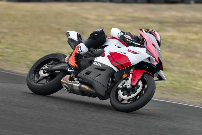 A rider in black and orange gear leans into a turn on a red and white Yamaha motorcycle on a racetrack, with blurred grass and tire barriers in the background.