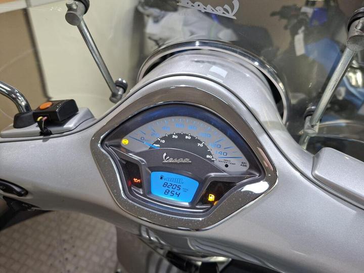 A close-up view of a Vespa scooter's dashboard, displaying a speedometer with a blue background, showing a speed of 0 mph and an odometer reading of 8205 miles. Various warning lights are illuminated, including indicators for engine and fuel.