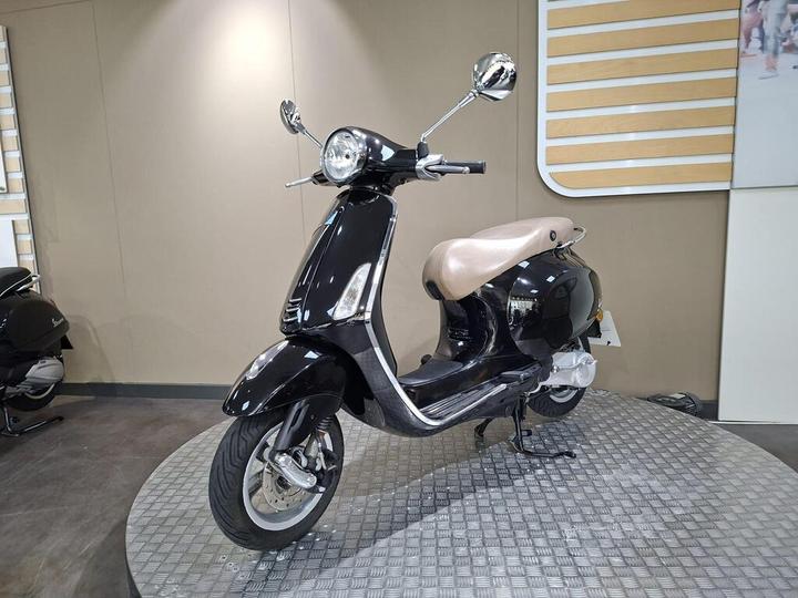 A sleek, black scooter with a retro design is showcased on a circular platform, featuring a light brown seat and chrome mirrors. The background is a plain wall with horizontal wooden slats.