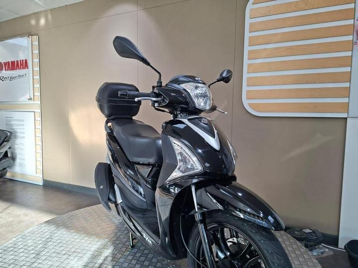 A black Yamaha scooter displayed indoors, featuring a sleek design, a protective windshield, and a rear storage box, with a light-colored wall and signage in the background.