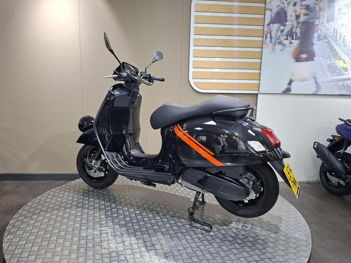 A black scooter with an orange stripe displayed on a circular platform in a showroom, with a blurred background of a busy street.