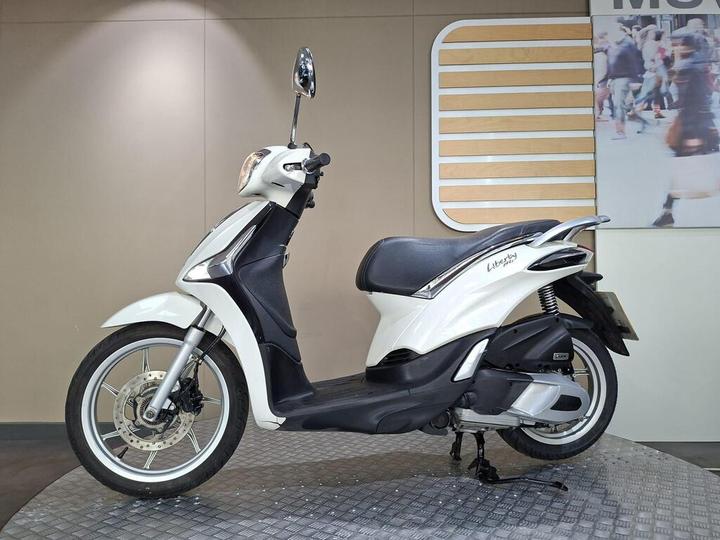 A white scooter is displayed against a neutral background, showcasing its sleek design, black seat, and chrome accents. The scooter is positioned on a circular metal platform, with a backdrop featuring a stylized graphic.