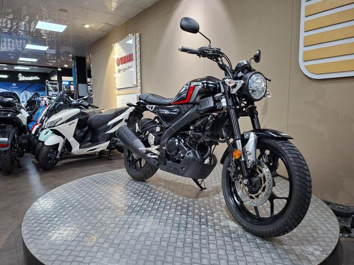 A black Yamaha motorcycle with a red accent is displayed on a circular platform in a showroom, surrounded by other bikes. The background features Yamaha branding and showroom lighting.