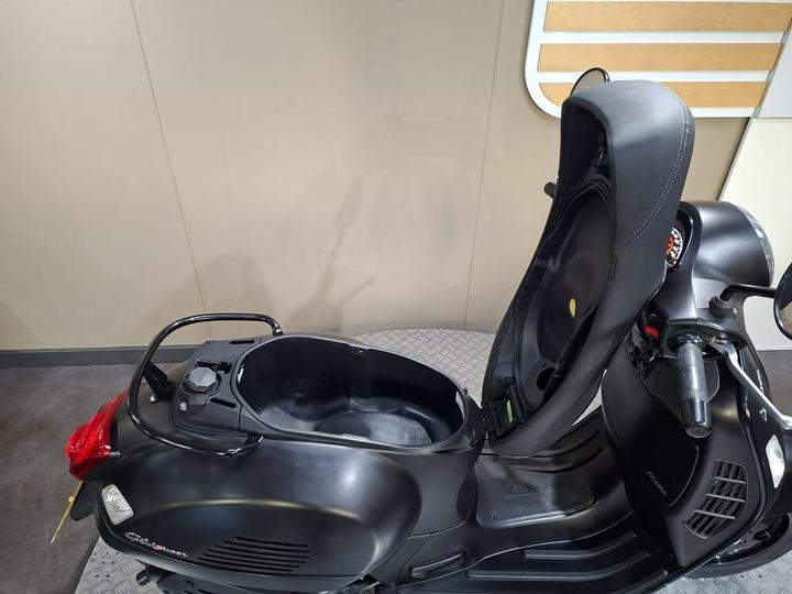 A black scooter with an open seat compartment, showcasing a spacious storage area beneath the seat, against a neutral background.