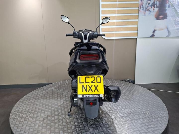 A black scooter viewed from the rear, displaying its license plate with the number "LC20 NXX," on a circular metal platform in a modern indoor setting.