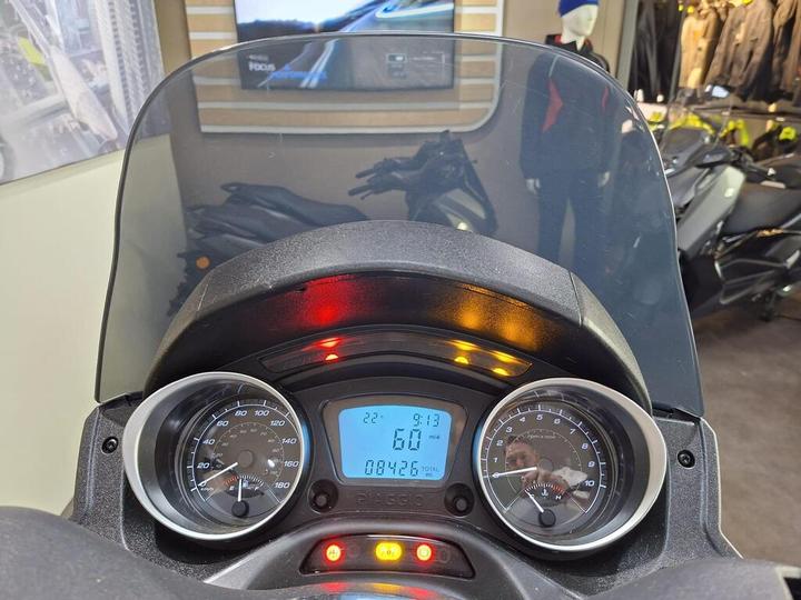 A close-up view of a scooter's dashboard, featuring a clear windshield, speedometer, fuel gauge, and a digital display showing the temperature at 22°C, time at 9:13, and total mileage of 08426 miles, with warning lights illuminated. The background includes scooters and riding gear in a showroom setting.