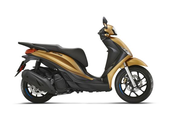 A sleek, modern scooter in a glossy gold finish, viewed from the side, showcasing its distinctive design features including a comfortable seat, stylized front headlight, and sporty wheels.