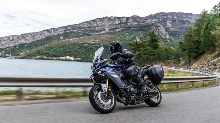 A motorcyclist in black gear navigates a winding road alongside a serene lake, with mountains and trees in the background.