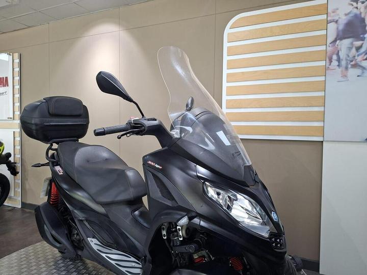 A black scooter with a windscreen and a rear storage compartment, positioned inside a showroom with neutral-colored walls and an abstract design panel in the background.