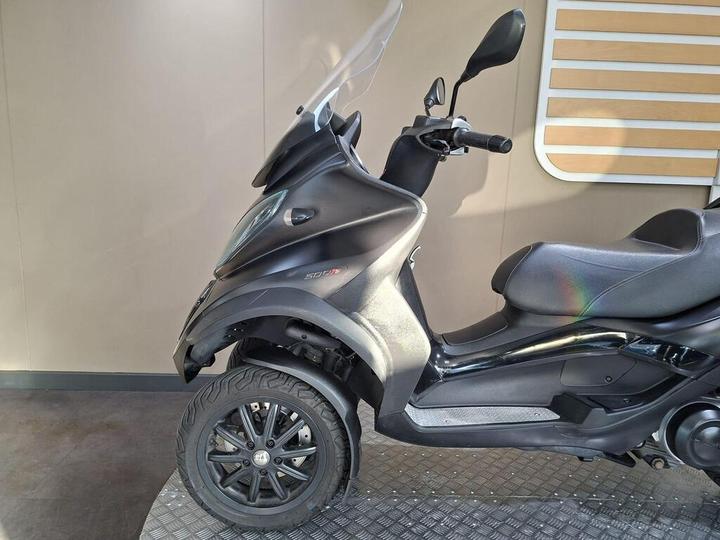 A side view of a three-wheeled scooter with a sleek black design, featuring a clear windscreen and a comfortable seat, on a textured floor against a simple background.