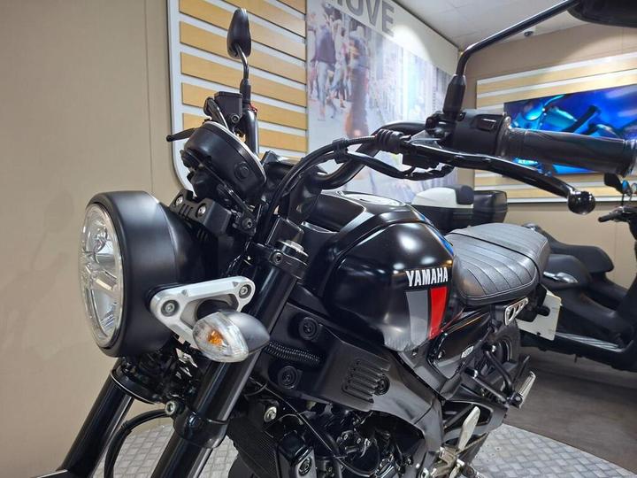 A close-up view of a black Yamaha motorcycle showcasing the front headlight, handlebars, and seat, set against a neutral background.