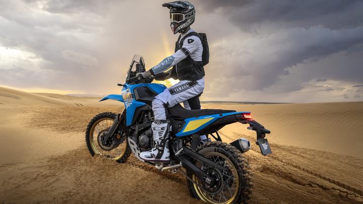 A motorcyclist dressed in black and white riding gear sits on a blue Yamaha Tenere 700 motorcycle in a desert landscape with rolling sand dunes and a cloudy sky.
