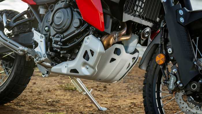 Close-up of a motorcycle engine showcasing a stainless steel skid plate, black frame components, and a rugged tire, positioned on a dirt surface.