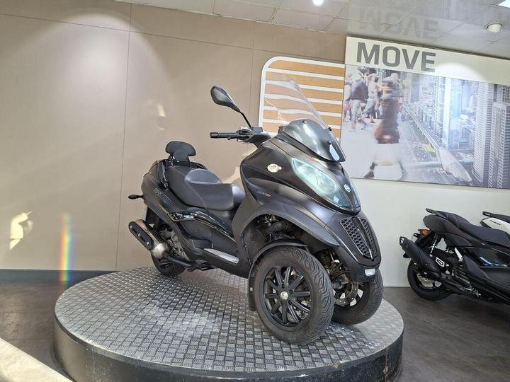 A black three-wheeled scooter displayed on a rotating platform in a showroom, with a modern cityscape mural in the background.