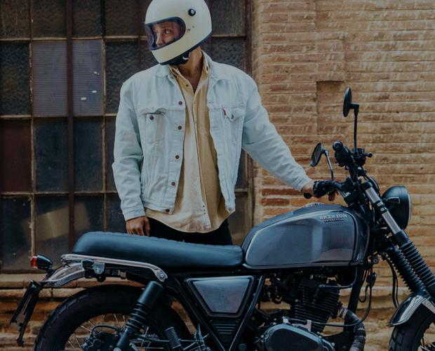 A man wearing a white helmet and blue corduroy jacket standing behind a classic blue motorcycle.