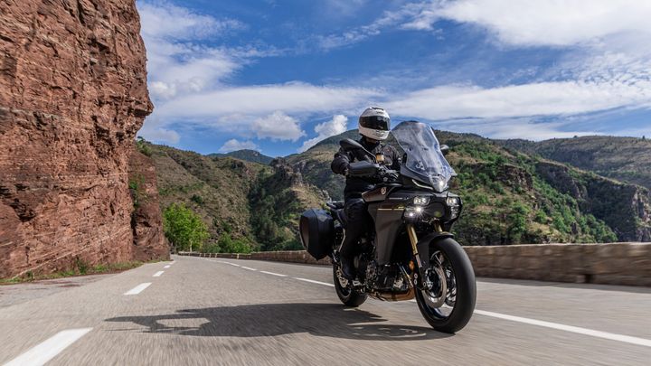 A motorcyclist in a black jacket and helmet rides along a winding road beside a steep, rocky cliff with green hills in the background under a blue sky with scattered clouds.