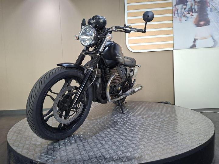 A black motorcycle displayed on a circular metallic platform, featuring a round headlight, rearview mirrors, and a modern design, against a neutral background.